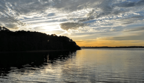 Sunset over a calm lake, with silhouetted trees and colorful clouds reflecting on the water's surface.