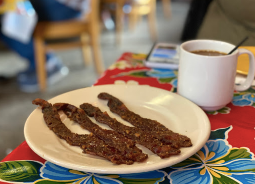 A plate with three strips of jerky next to a white mug of coffee on a colorful floral tablecloth.