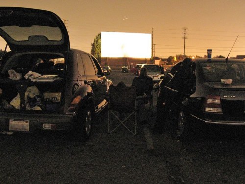 A drive-in movie scene at dusk, with two cars parked and people sitting outside watching a large screen.
