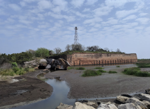 A weathered brick structure surrounded by greenery and water, with a tower in the background under a cloudy sky.