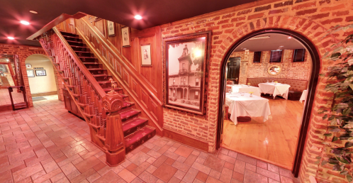 A warmly lit restaurant interior featuring a wooden staircase, brick walls, and dining tables visible through an archway.