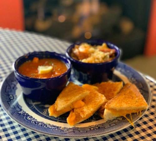 A plate with grilled cheese sandwiches, soup, and a side dish, set on a checkered tablecloth with a warm background.