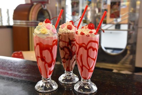 Three colorful milkshakes in tall glasses, topped with whipped cream, cherries, and candy, on a black countertop.