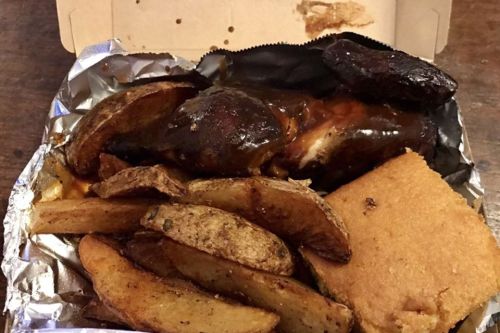 A takeout box containing BBQ chicken, seasoned potato wedges, and a piece of cornbread.