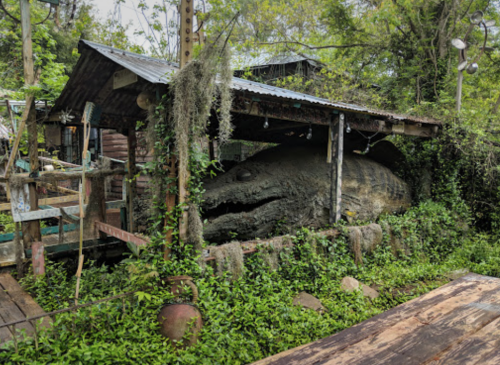 A large, moss-covered sculpture of a fish is nestled in a lush, green garden under a wooden shelter.