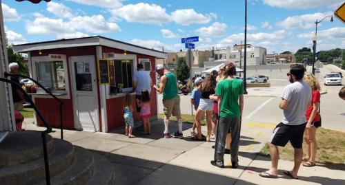 A busy outdoor food stand with a line of people waiting, surrounded by a sunny park and city buildings in the background.
