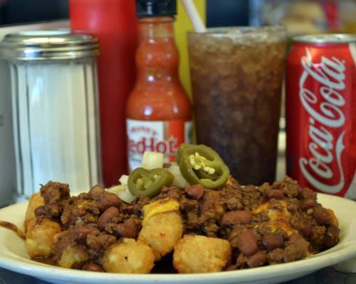 A plate of tater tots topped with chili, beans, jalapeños, and condiments, with a soda and hot sauce in the background.