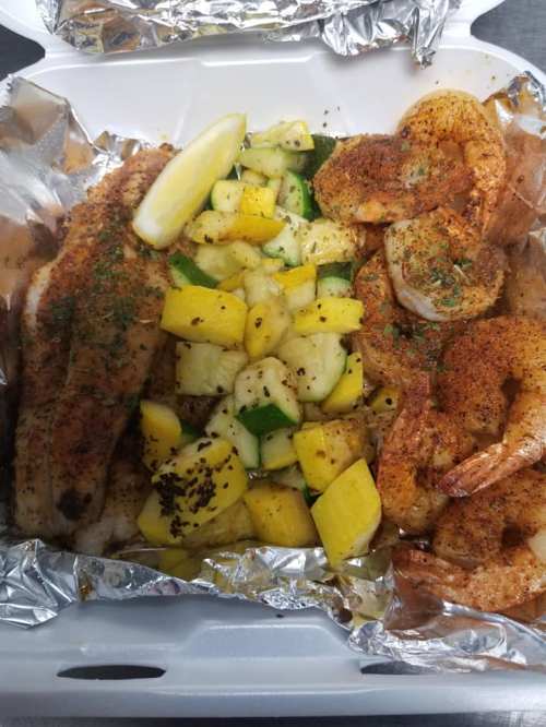 A takeout container with seasoned shrimp, fish, and a side of grilled vegetables and lemon wedges.