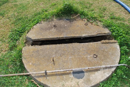 A round, concrete manhole cover partially open, surrounded by grass and a metal pipe nearby.