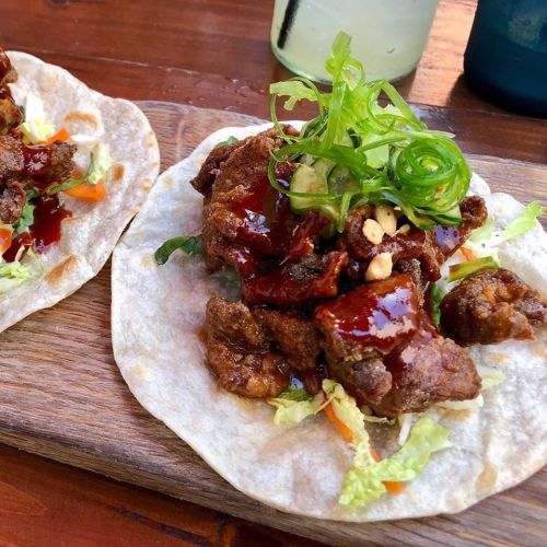 Two soft tacos filled with crispy meat, topped with green onions, peanuts, and a drizzle of sauce, served on a wooden board.