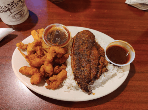 A plate of seasoned fish fillet on rice, accompanied by fried shrimp and a small container of sauce.