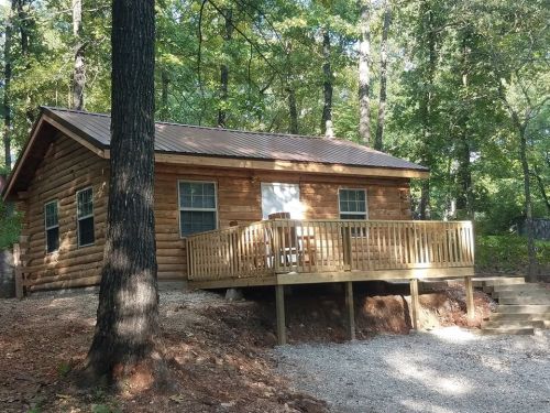 A rustic log cabin with a wooden deck, surrounded by trees and gravel, set in a peaceful outdoor environment.