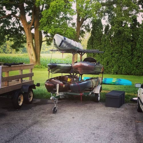 A trailer with stacked kayaks and a paddleboard parked on a gravel surface, surrounded by trees and greenery.