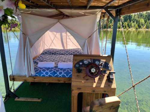 A cozy floating bed with a steering wheel, surrounded by calm water and greenery, under a canopy.