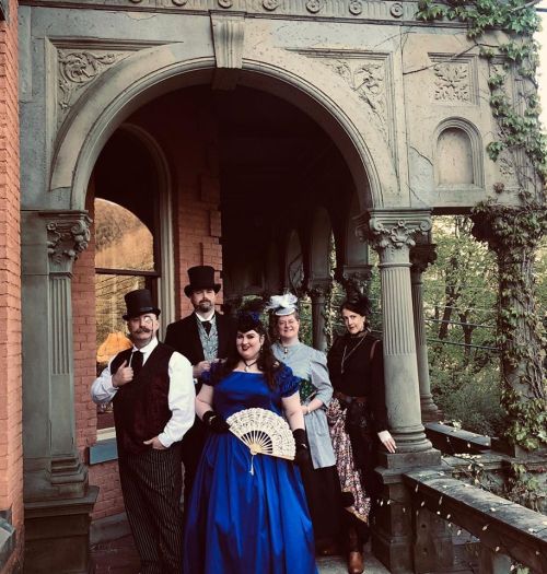A group of five people in vintage attire poses on a grand porch with ornate architecture and greenery.