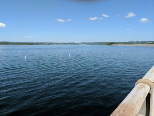 A calm lake under a clear blue sky, with a wooden railing in the foreground and distant hills on the horizon.