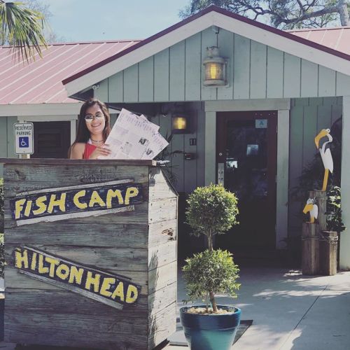 A woman stands at the entrance of Fish Camp in Hilton Head, holding menus, with a decorative pelican nearby.
