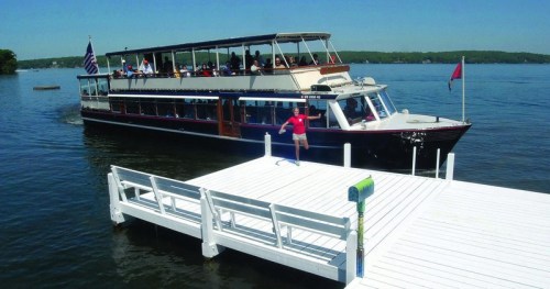 A large boat approaches a white dock on a sunny day, with passengers visible on board and a person preparing to board.