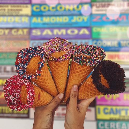 A hand holds five ice cream cones, each topped with colorful sprinkles and chocolate, against a vibrant menu backdrop.