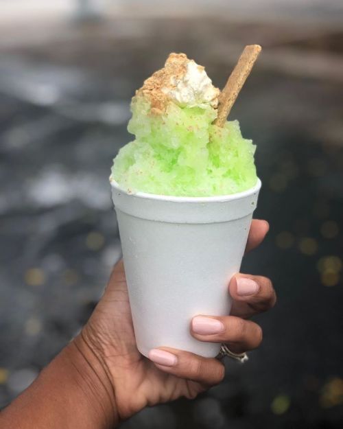 A hand holding a white cup filled with green shaved ice, topped with whipped cream and a cookie.