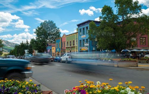 Colorful buildings line a street with blooming flowers in the foreground and clouds in a blue sky above.