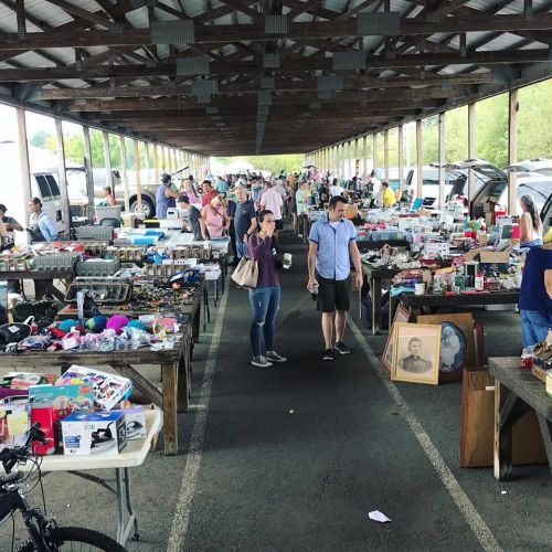 A bustling outdoor market with people browsing tables filled with various items under a covered structure.