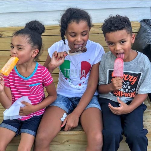 Three children sitting on a bench, enjoying colorful popsicles on a sunny day.