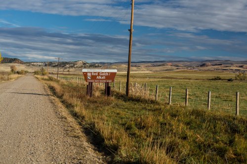 A dirt road leads to a sign for Red Gulch, with rolling hills and a cloudy sky in the background.