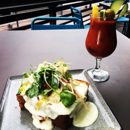 A plate of toast topped with creamy sauce and fresh greens, alongside a colorful cocktail on a table.