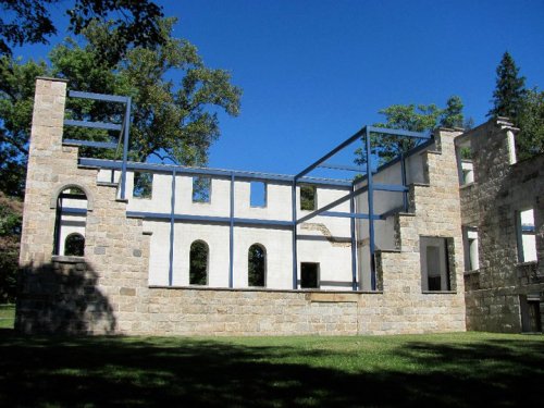 Ruins of a stone building with a metal frame, set against a clear blue sky and surrounded by trees.