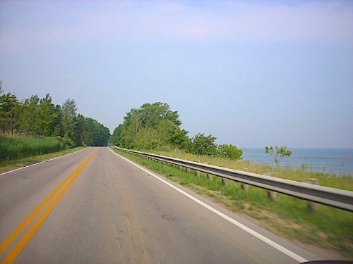 A scenic road lined with greenery, running parallel to a calm body of water under a clear blue sky.