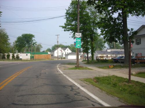 A rural road with a highway sign for Route 531, lined with trees and houses in the background.