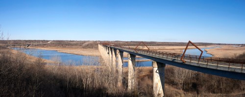 A long bridge spans a river, surrounded by bare trees and open fields under a clear blue sky.