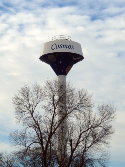 A water tower painted with "Cosmos," featuring celestial designs, stands against a cloudy sky and bare trees.