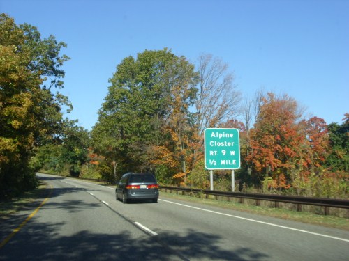 A car drives on a road with a green sign indicating Alpine Closter, RT 9 W, ½ mile ahead, surrounded by autumn trees.