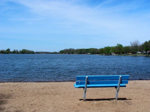 A blue bench on a sandy shore overlooking a calm lake under a clear blue sky.