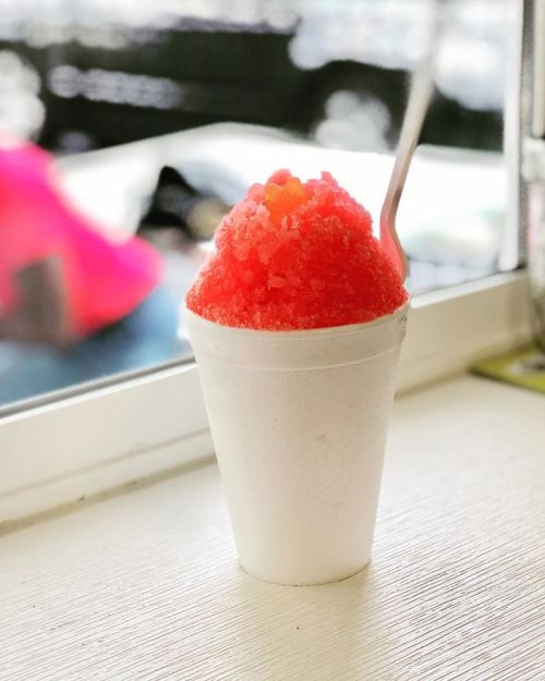 A white cup filled with bright red shaved ice, topped with colorful sprinkles, sits on a table by a window.