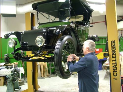 A mechanic works on a vintage black car lifted on a hoist in a workshop. Tools and equipment are visible in the background.
