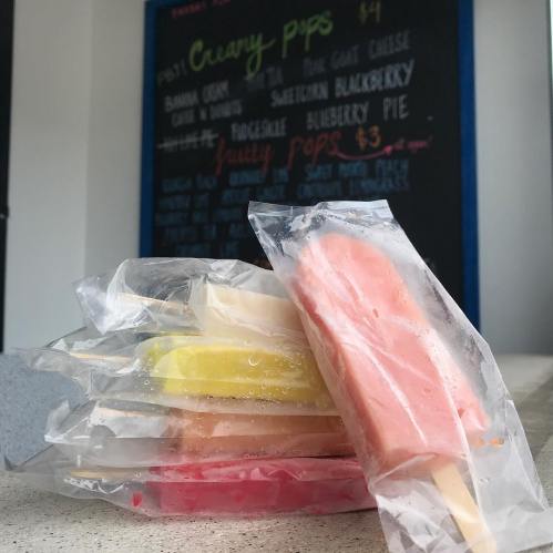 A stack of colorful popsicles in clear wrappers, with a chalkboard menu of flavors in the background.