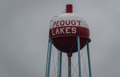 Water tower with "PEQUOT LAKES" written in bold red letters against a gray sky.