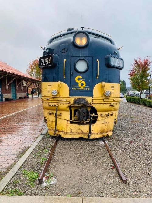 Front view of a vintage blue and yellow train engine on tracks, with a station and autumn trees in the background.