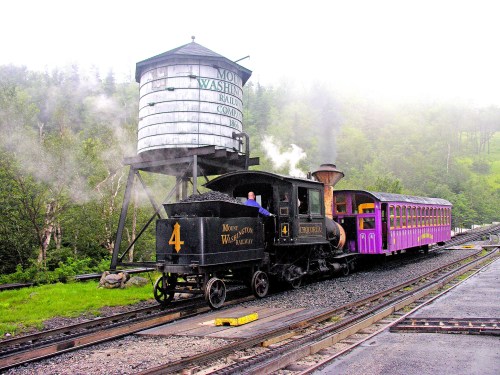 A vintage steam train with a purple passenger car stops near a water tower in a misty, green landscape.