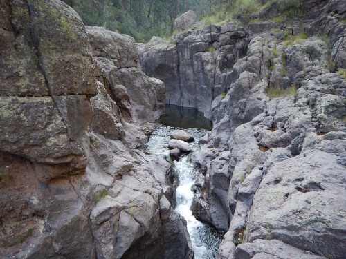 A rocky landscape with a small waterfall flowing through a narrow canyon, surrounded by greenery and steep cliffs.