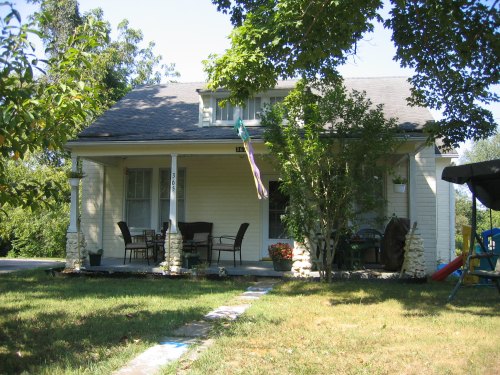A charming two-story house with a porch, surrounded by trees and a grassy yard, featuring outdoor seating and colorful decor.