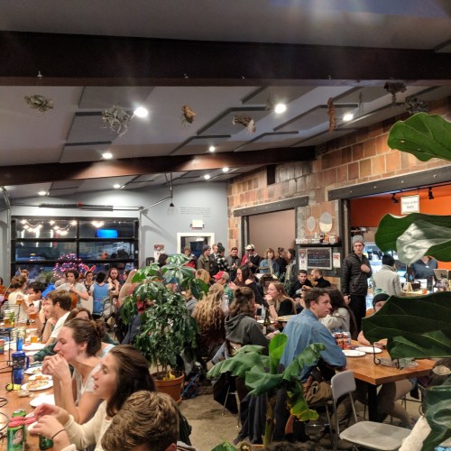 A busy restaurant interior with people dining, plants, and a lively atmosphere.