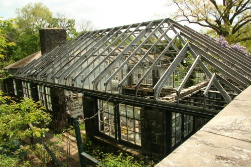 A partially constructed greenhouse with a metal frame, surrounded by greenery and trees under a bright sky.