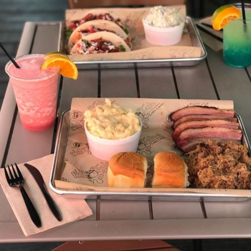 A table with trays of food: tacos, macaroni, pulled pork, sliders, and colorful drinks.
