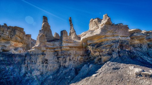 Rock formations with unique shapes under a clear blue sky, showcasing layers of sediment and a hint of the moon.