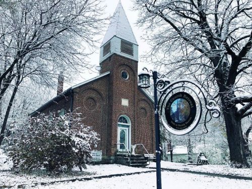 A snow-covered church with a tall steeple, surrounded by trees and a decorative sign in front.