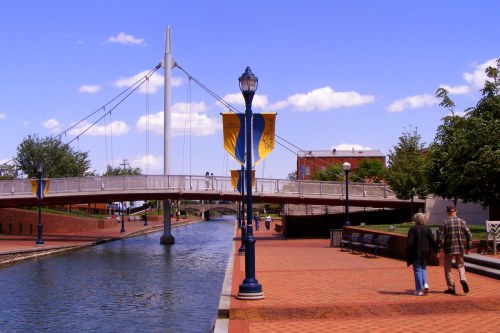 A scenic walkway along a canal, featuring a bridge, lampposts, and people strolling under a blue sky with clouds.
